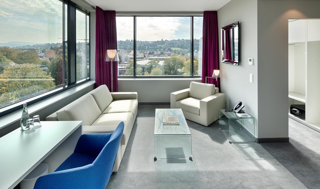 Modern suite with large windows, sofa, armchair, desk and city view. Light colors and stylish furnishings dominate the room., &copy; Hotel am Remspark