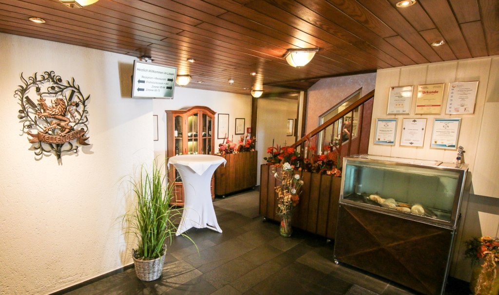 Eingangsbereich mit Holzwand, Dekorationen, Aquarium und einem Wappen. Ein Tisch mit weißer Tischdecke steht neben Pflanzen., © Ulli Kellenbenz Eingangsbereich mit Holzwand, Dekorationen, Aquarium und einem Wappen. Ein Tisch mit weißer Tischdecke steht neben Pflanzen., © Ulli Kellenbenz