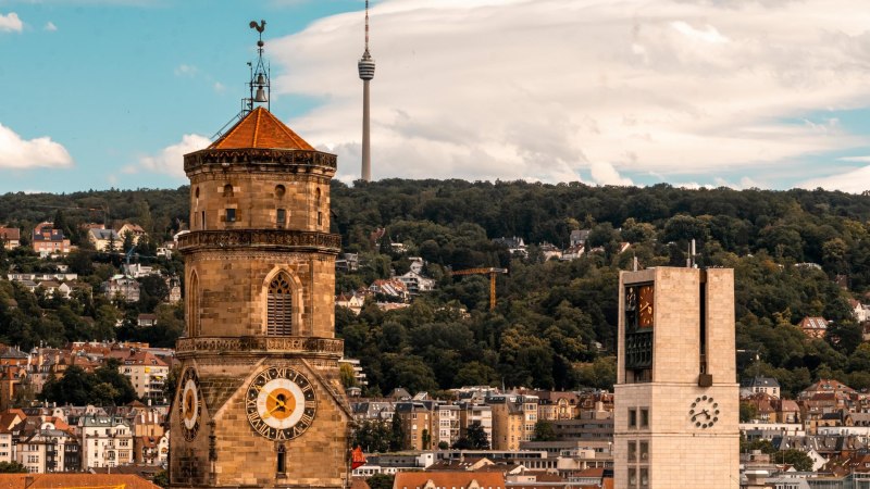 Die Stiftskirche in Stuttgart mit dem markanten Turm und der Uhr im Vordergrund. Rechts im Bild der Rathausturm. Im Hintergrund ist der Fernsehturm zu sehen, umgeben von gr&uuml;nen H&uuml;geln., &copy; Stuttgart-Marketing GmbH, Sarah Schmid