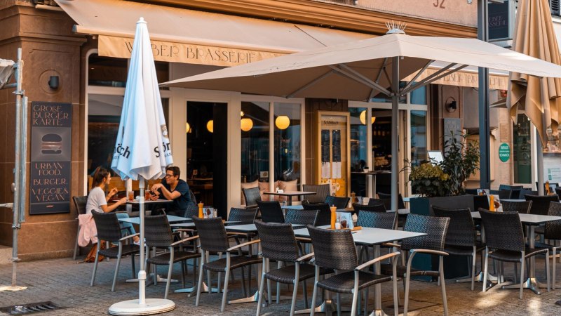 Ein Café mit Terrasse, Tischen und Stühlen, Sonnenschirmen und Gästen. Ein Schild wirbt für Frühstücksangebote., © Stuttgart Marketing GmbH, Sarah Schmid Ein Café mit Terrasse, Tischen und Stühlen, Sonnenschirmen und Gästen. Ein Schild wirbt für Frühstücksangebote., © Stuttgart Marketing GmbH, Sarah Schmid