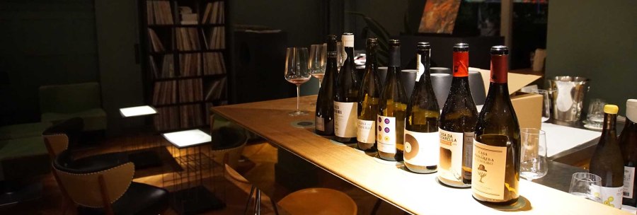 Several bottles of wine stand on a wooden table in a stylish bar. A record shelf and comfortable seating can be seen in the background., &copy; Weinhandlung Kreis KG