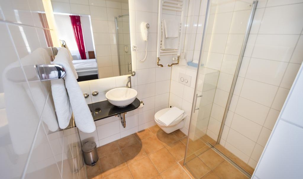 Modernes Badezimmer mit Glasdusche, Waschbecken, Handtüchern und Toilette. Weiße Fliesen und ein Spiegel sind sichtbar., © ARNOTEL GmbH