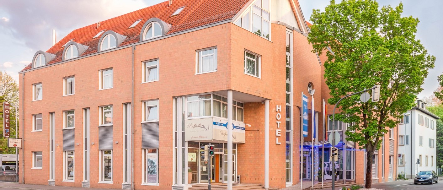 Modernes Hotelgeb&auml;ude mit rotem Ziegeldach und gro&szlig;en Fenstern an einer Stra&szlig;enecke, umgeben von B&auml;umen., &copy; TOMAS