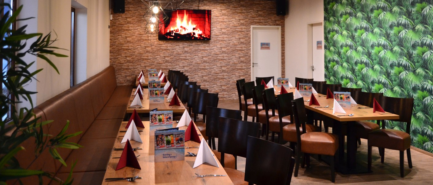 Ein langer Tisch in einer Lounge, dekoriert mit roten und wei&szlig;en Servietten. An der Wand ein Bildschirm mit Kaminfeuer, daneben eine gr&uuml;ne Tapete., &copy; Bowling & Billard Lounge