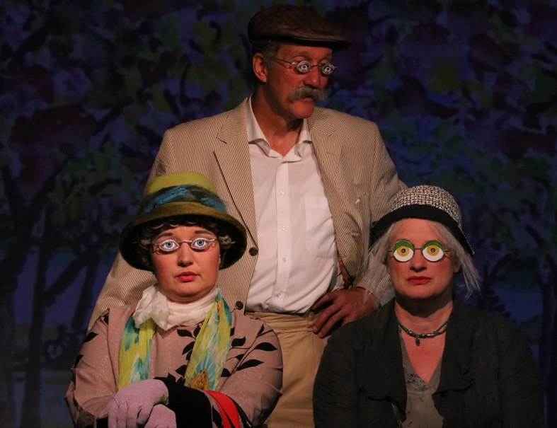 Three people in colorful costumes and funny glasses stand on a stage. The background shows a colorful, abstract landscape., &copy; @Foto_Katja Ritter