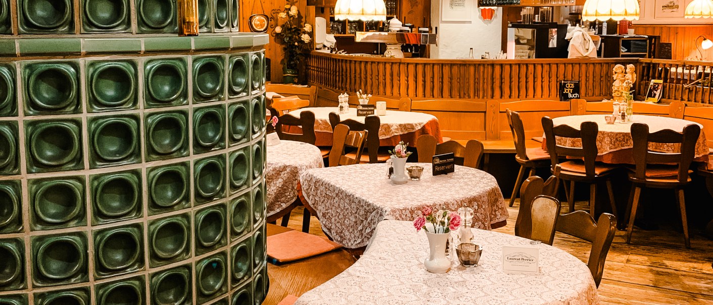 Gemütliches Restaurant mit Holzmöbeln, Tischdecken und einem grünen Kachelofen. Lampen hängen von der Decke, Bar im Hintergrund., © Weinstube Kachelofen Gemütliches Restaurant mit Holzmöbeln, Tischdecken und einem grünen Kachelofen. Lampen hängen von der Decke, Bar im Hintergrund., © Weinstube Kachelofen