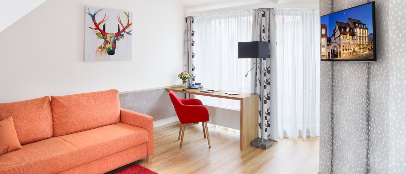 Helle Junior Suite mit oranger Couch, rotem Stuhl, Schreibtisch, Wandkunst und TV. Große Fenster mit Vorhängen, Holzboden und moderner Einrichtung., © Hirsch Hotel