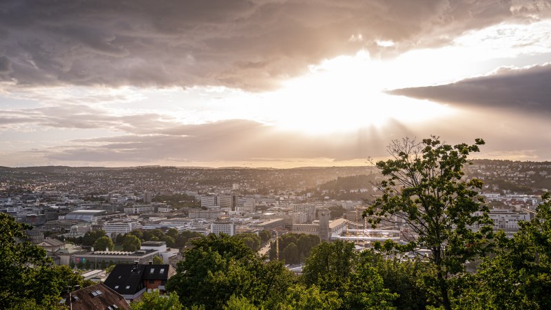 Sonnenuntergang über Stuttgart von der Uhlandshöhe aus. Die Stadt liegt unter einem bewölkten Himmel, durch den Sonnenstrahlen brechen., © Christine Garcia Urbina trickytine Sonnenuntergang über Stuttgart von der Uhlandshöhe aus. Die Stadt liegt unter einem bewölkten Himmel, durch den Sonnenstrahlen brechen., © Christine Garcia Urbina trickytine