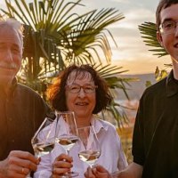 Drei Personen sto&szlig;en mit Weingl&auml;sern an, umgeben von Palmen im Sonnenuntergang., &copy; Weingut Bayer