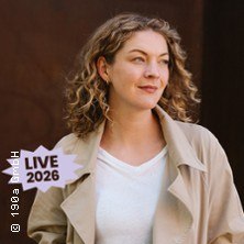 Eine Frau mit lockigem Haar tr&auml;gt einen beigen Mantel und schaut zur Seite. Der Schriftzug "LIVE 2026" ist im Bild., &copy; links im Bild