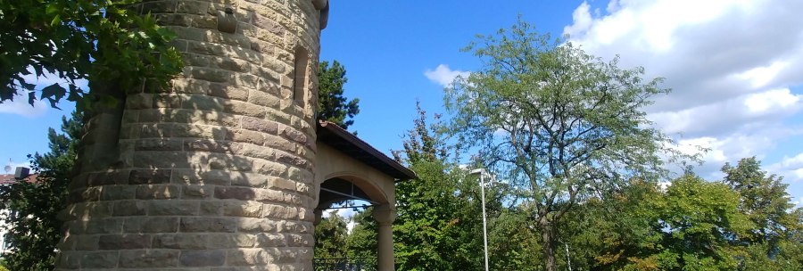 Runder Steinturm mit Zinnen, umgeben von B&auml;umen und blauem Himmel. Der Turm steht auf einer gepflasterten Fl&auml;che., &copy; SMG