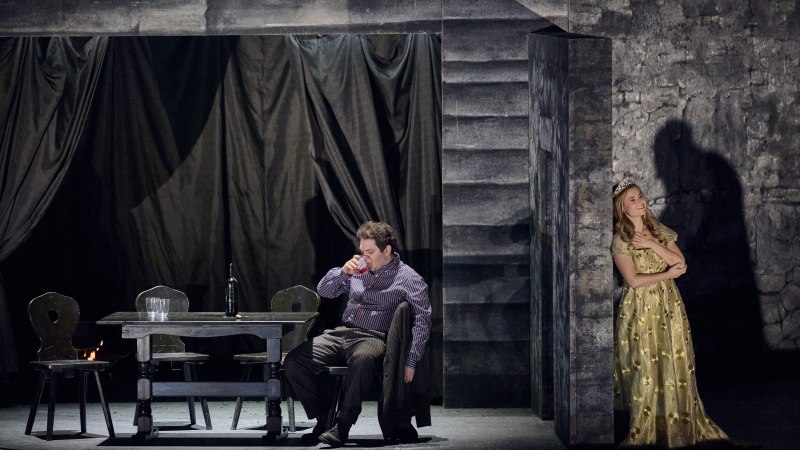 Ein Mann sitzt an einem Tisch und trinkt, während eine Frau in einem gelben Kleid an der Wand lehnt. Die Bühne ist dunkel und dramatisch beleuchtet., © Martin Sigmund Ein Mann sitzt an einem Tisch und trinkt, während eine Frau in einem gelben Kleid an der Wand lehnt. Die Bühne ist dunkel und dramatisch beleuchtet., © Martin Sigmund