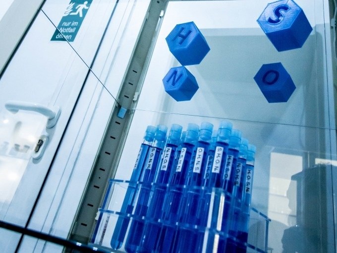 Blue illuminated test tubes on a shelf, above chemical symbols H, N, S, O. Emergency exit sign in the background., © ExitGames Stuttgart Blue illuminated test tubes on a shelf, above chemical symbols H, N, S, O. Emergency exit sign in the background., © ExitGames Stuttgart