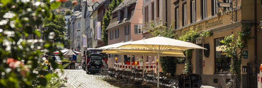 Cobbled street in the Bohnenviertel with Weinhaus Stetter, outdoor seating area under parasols, surrounded by flowering plants and historic buildings., © SMG Stuttgart Marketing GmbH - Sarah Schmid