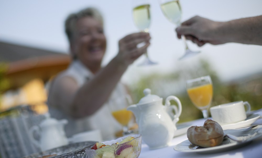 Two people are clinking their champagne glasses at an outdoor table. There is fruit, rolls, and orange juice on the table., © Badhotel Restaurant Stauferland Two people are clinking their champagne glasses at an outdoor table. There is fruit, rolls, and orange juice on the table., © Badhotel Restaurant Stauferland