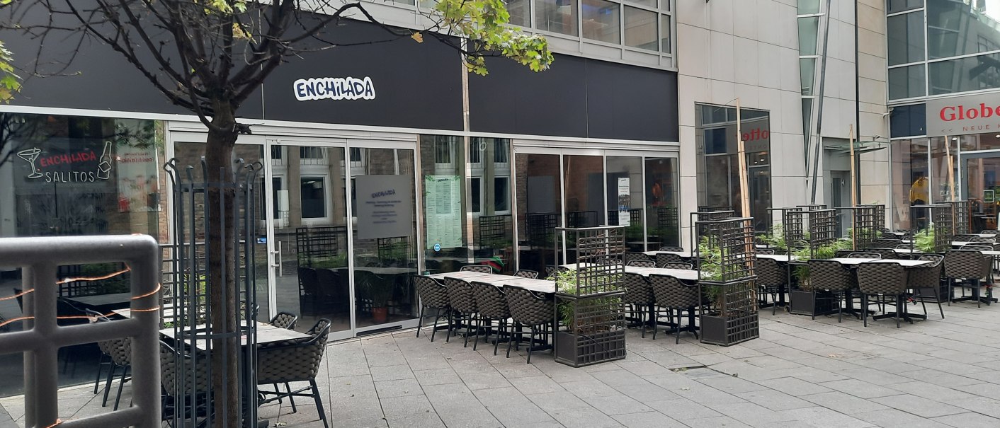 Outdoor area of a restaurant called Enchilada with empty tables and chairs. A tree and plants separate the seating areas., &copy; SMG