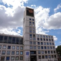 A modern building with a tall clock tower and lots of windows. The sky is blue with a few clouds., © Fei Shiyu
