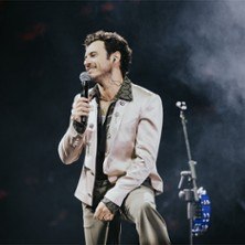 Ein Sänger in einem eleganten Anzug hält ein Mikrofon auf einer Bühne. Im Hintergrund ist ein Ständer mit einem Tamburin zu sehen., © links im Bild