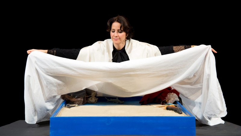 Eine Frau in einem schwarzen Outfit hält ein weißes Tuch über eine blaue Kiste mit Sand. Darin liegen verschiedene Objekte, darunter eine Pistole., © Gubás Gabi Eine Frau in einem schwarzen Outfit hält ein weißes Tuch über eine blaue Kiste mit Sand. Darin liegen verschiedene Objekte, darunter eine Pistole., © Gubás Gabi