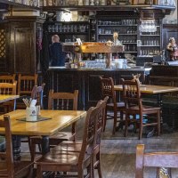 Gemütlicher Innenraum eines Restaurants mit Holztischen und -stühlen, einer Bar im Hintergrund und einem großen Kachelofen links., © SMG, Sarah Schmid Gemütlicher Innenraum eines Restaurants mit Holztischen und -stühlen, einer Bar im Hintergrund und einem großen Kachelofen links., © SMG, Sarah Schmid