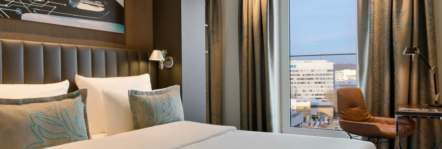 Modernes Hotelzimmer mit großem Bett, Kunstwerk an der Wand, Fenster mit Stadtblick, braunem Sessel und Schreibtisch., © TOMAS Modernes Hotelzimmer mit großem Bett, Kunstwerk an der Wand, Fenster mit Stadtblick, braunem Sessel und Schreibtisch., © TOMAS