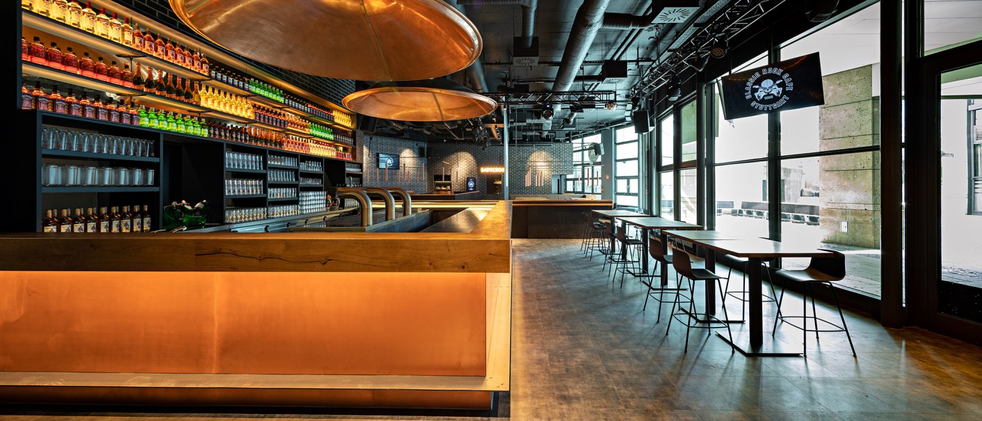 Modernes Café mit Bar, beleuchteten Regalen voller Flaschen, Gläsern und Sitzbereichen. Große Fenster lassen viel Licht herein., © Maks Richter