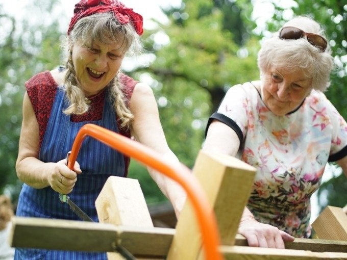 Two older women are sawing wood together outdoors. They look cheerful and focused. One is wearing a headscarf, the other sunglasses., &copy; Cool-Tours StattReisen
