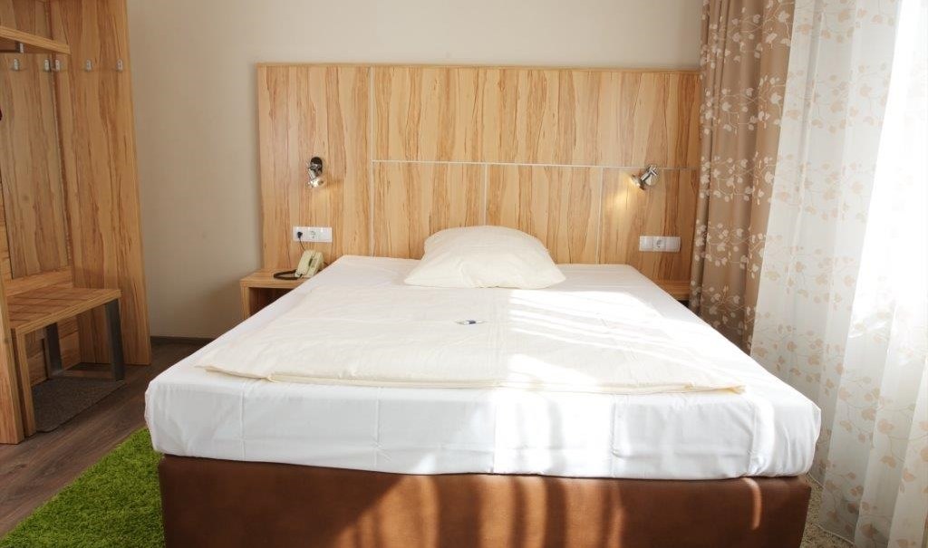 A cozy single room with a bed, wooden furniture and floral curtains. There is a green carpet on the floor., © Hotel Reinhardtshof Garni A cozy single room with a bed, wooden furniture and floral curtains. There is a green carpet on the floor., © Hotel Reinhardtshof Garni