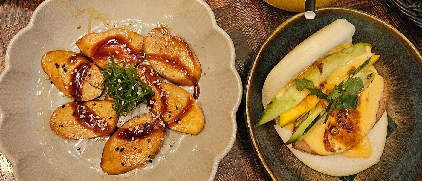 A plate of vegan dumplings in sauce, a bao bun with vegetables and a drink with ice cubes on a wooden table., © SMG