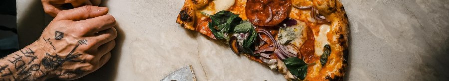 A half-eaten pizza with basil and onions on a light-colored stone, with a pizza cutter and scissors next to it; on the left, two tattooed hands., &copy; L'Artista, Stuttgart
