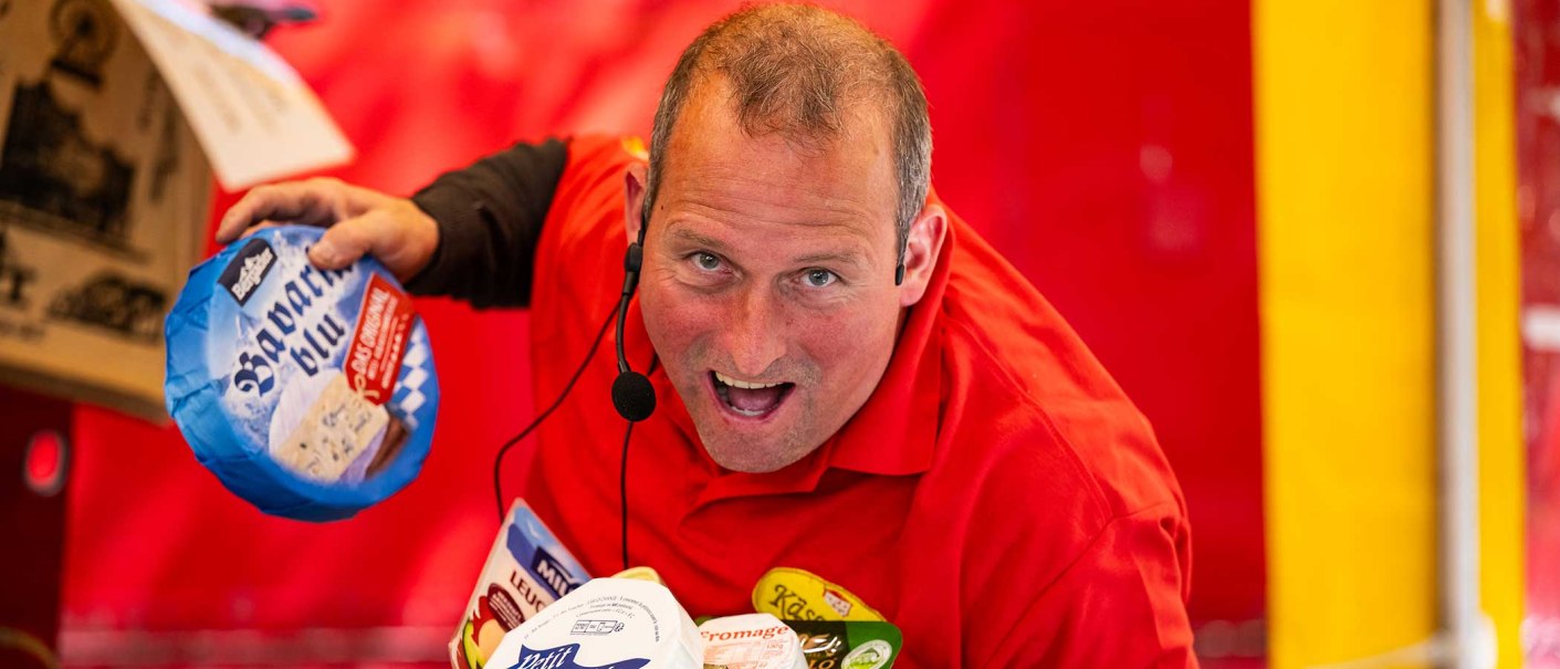Ein Verkäufer in rotem Hemd mit Mikrofon präsentiert Käseprodukte auf einem Marktstand. Hintergrund in Rot und Gelb., © Tom Weller Ein Verkäufer in rotem Hemd mit Mikrofon präsentiert Käseprodukte auf einem Marktstand. Hintergrund in Rot und Gelb., © Tom Weller