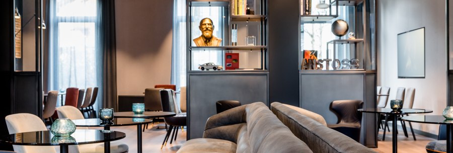 Moderne Lounge mit grauen Sofas, runden Tischen und dekorativen Elementen wie einer Büste und Büchern in Regalen., © TOMAS Moderne Lounge mit grauen Sofas, runden Tischen und dekorativen Elementen wie einer Büste und Büchern in Regalen., © TOMAS