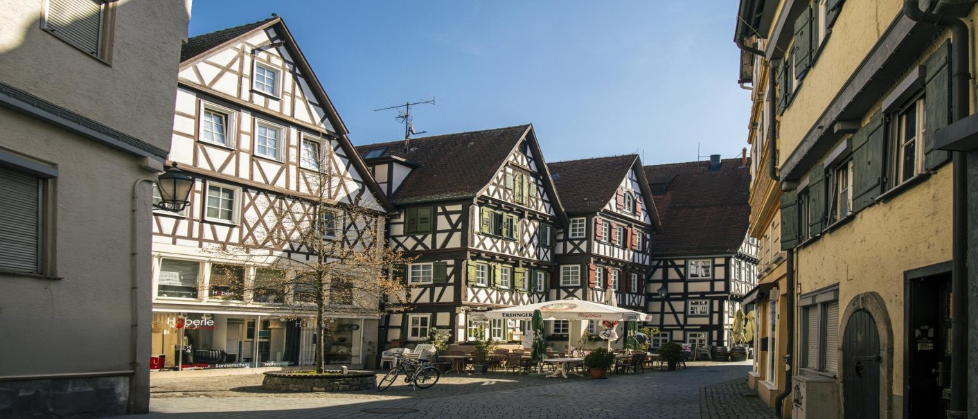 Fachwerkhäuser am Marktplatz in Schorndorf, sonniger Tag, leere Straße, ein Fahrrad steht vor einem Café mit Sonnenschirmen., © SMG, Sarah Schmid Fachwerkhäuser am Marktplatz in Schorndorf, sonniger Tag, leere Straße, ein Fahrrad steht vor einem Café mit Sonnenschirmen., © SMG, Sarah Schmid