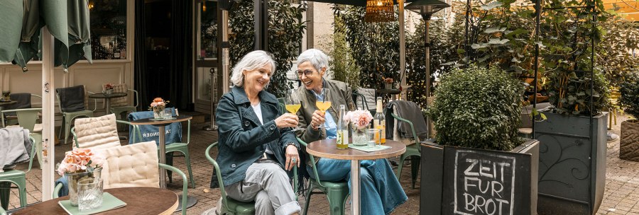 Zwei Frauen sitzen in einem gem&uuml;tlichen Caf&eacute; im Freien, sto&szlig;en mit Getr&auml;nken an und lachen. Ein Schild bietet Zimtschnecken an., &copy; Stuttgart Marketing GmbH - Sarah Schmid