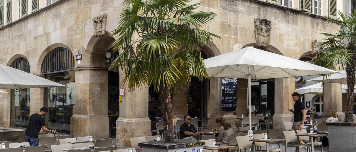 Café with tables and chairs under parasols in front of a historic building with arcades. Palm trees and people in the outdoor area., © SMG, Sarah Schmid