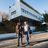 Two women walk in front of the Weissenhof Museum in Stuttgart. The building by Le Corbusier is modern and white, with clear lines and large windows., &copy; SMG, WP Steinheisser