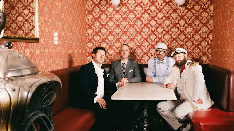 Vier Männer in eleganter Kleidung sitzen an einem Tisch in einem Raum mit roter, gemusterter Tapete und antikem Ofen., © Theaterhaus Stuttgart e.V.