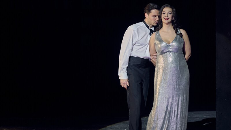 Ein Mann im Smoking und eine Frau im silbernen Abendkleid stehen eng beieinander auf einer Bühne, in dramatischer Beleuchtung., © Württembergische Staatstheater Stuttgart Ein Mann im Smoking und eine Frau im silbernen Abendkleid stehen eng beieinander auf einer Bühne, in dramatischer Beleuchtung., © Württembergische Staatstheater Stuttgart