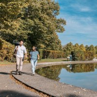 A couple is walking in Wartberg Park along a pond, with a large sculpture and a modern building in the background., © Stuttgart-Marketing GmbH, Sarah Schmid A couple is walking in Wartberg Park along a pond, with a large sculpture and a modern building in the background., © Stuttgart-Marketing GmbH, Sarah Schmid