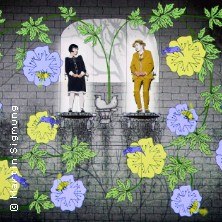 Zwei Figuren stehen auf Podesten vor einem gemauerten Hintergrund, umgeben von großen, bunten Blumen. Die Szene wirkt märchenhaft und fantasievoll., © links im Bild Zwei Figuren stehen auf Podesten vor einem gemauerten Hintergrund, umgeben von großen, bunten Blumen. Die Szene wirkt märchenhaft und fantasievoll., © links im Bild