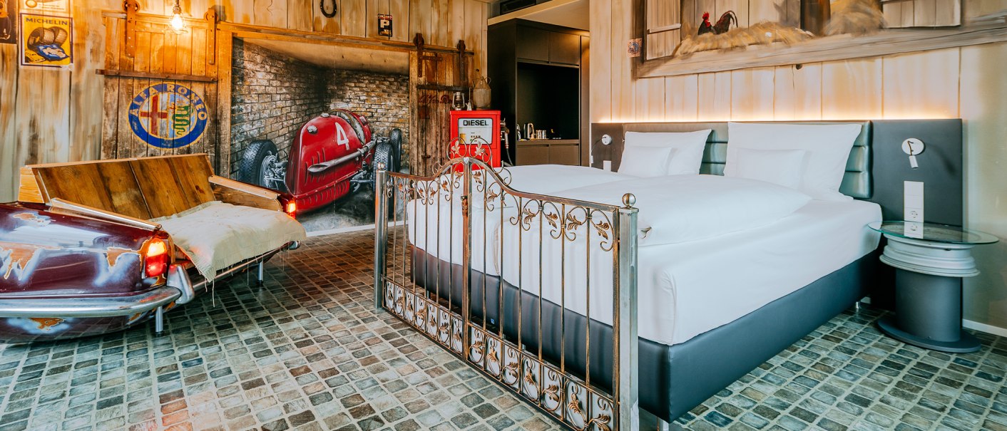Themenzimmer im V8 Hotel mit einem Bett, Autodekorationen, einer alten Zapfsäule und einem Sofa aus einem Autoteil. Wände mit Rennwagenmotiv., © Thomas Niedermüller Themenzimmer im V8 Hotel mit einem Bett, Autodekorationen, einer alten Zapfsäule und einem Sofa aus einem Autoteil. Wände mit Rennwagenmotiv., © Thomas Niedermüller