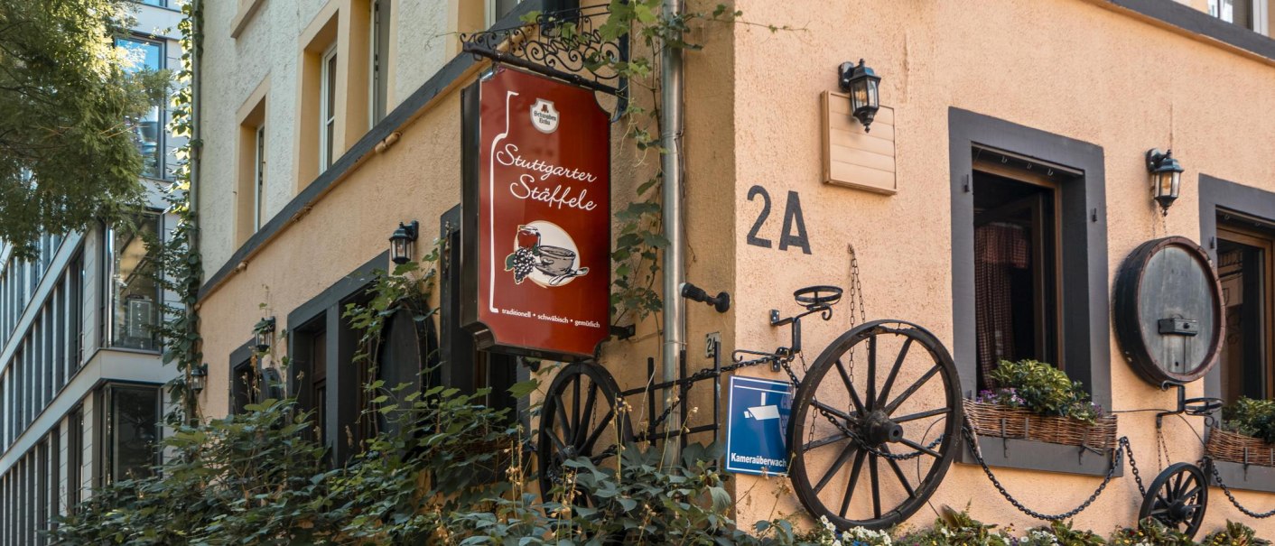 Building with the sign 'Stuttgarter Stäffele', decorated with wagon wheels and plants. The façade is beige with black window frames., © SMG Stuttgart Marketing GmbH - Sarah Schmid Building with the sign 'Stuttgarter Stäffele', decorated with wagon wheels and plants. The façade is beige with black window frames., © SMG Stuttgart Marketing GmbH - Sarah Schmid