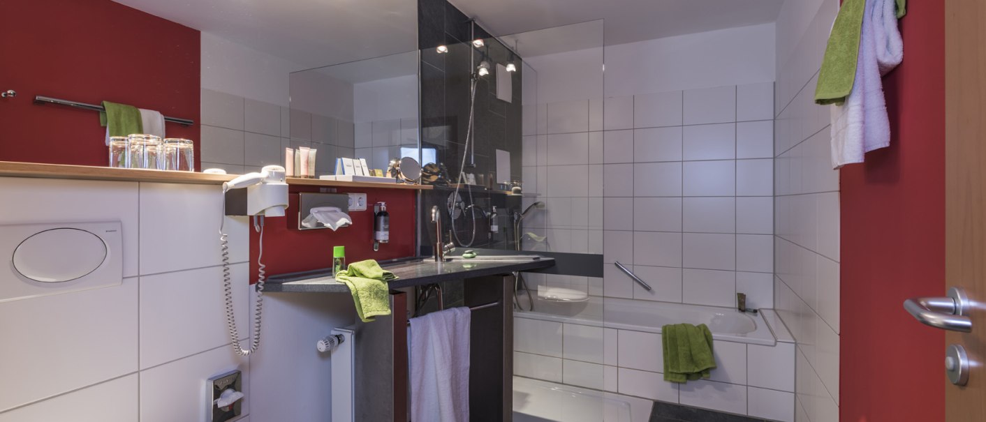 Modernes Badezimmer mit roten Akzentwänden, Dusche, Badewanne, Waschbecken und Handtüchern. Beleuchtung an der Decke, Fliesenboden., © Hotel Berlin aZIS Hotel BetriebsGmbH