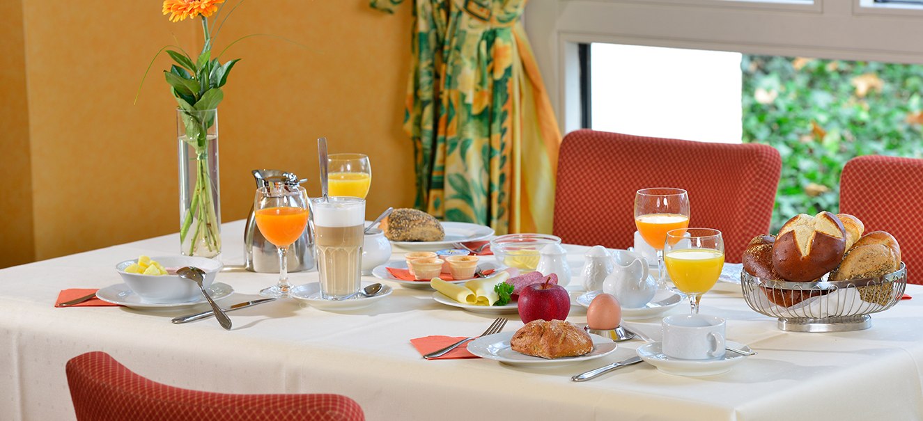 A breakfast table with bread rolls, juice, coffee, an egg, fruit and a flower in a vase, surrounded by red chairs., © Amber Hotel Leonberg / Stuttgart A breakfast table with bread rolls, juice, coffee, an egg, fruit and a flower in a vase, surrounded by red chairs., © Amber Hotel Leonberg / Stuttgart