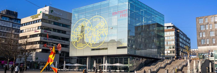 The Stuttgart Art Museum with its striking glass cube on Schlossplatz. A colorful sculpture stands in the foreground, surrounded by modern buildings., © Stuttgart-Marketing GmbH, Sarah Schmid