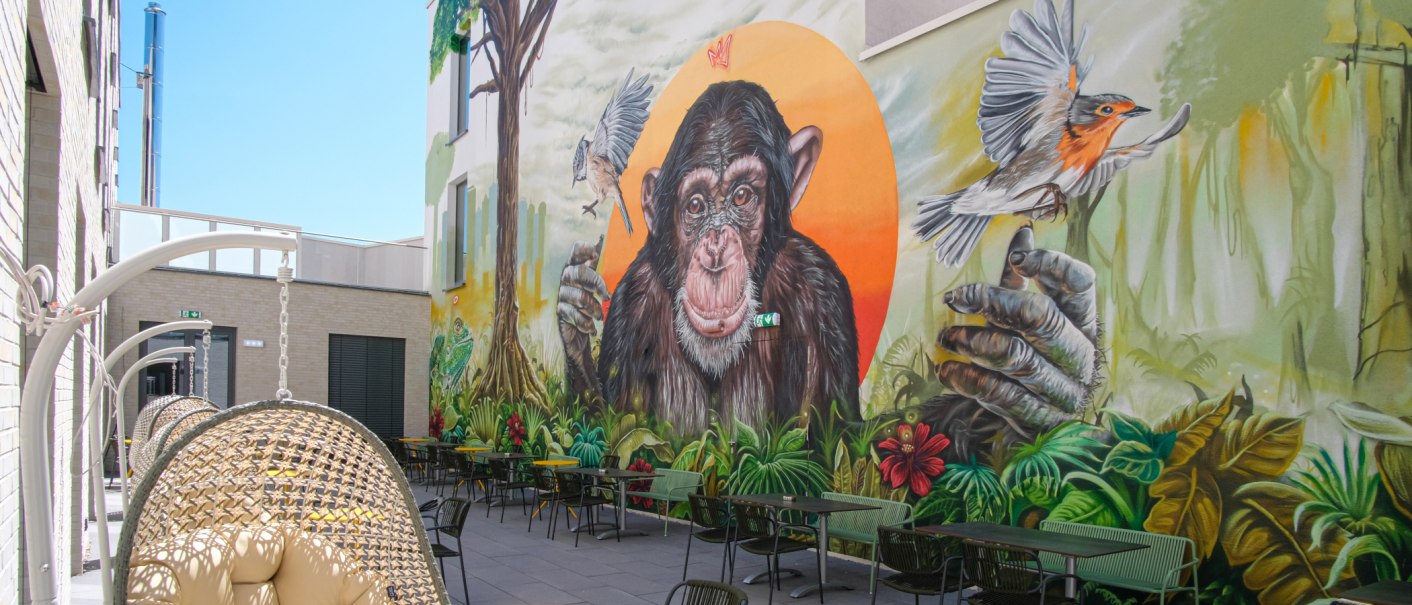 Terrasse mit H&auml;ngesesseln und Tischen, Wandgem&auml;lde zeigt einen Schimpansen und V&ouml;gel vor einem orangefarbenen Hintergrund., &copy; 13 6 Design GmbH (Judenhof 10, 89073 Ulm)