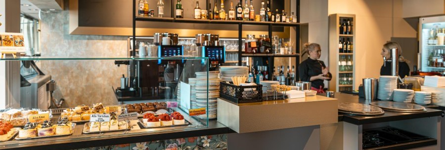 A caf&eacute; with a counter, cakes in a display case and shelves with bottles in the background. Two people are standing behind the counter., &copy; SMG Stuttgart Marketing GmbH - Sarah Schmid