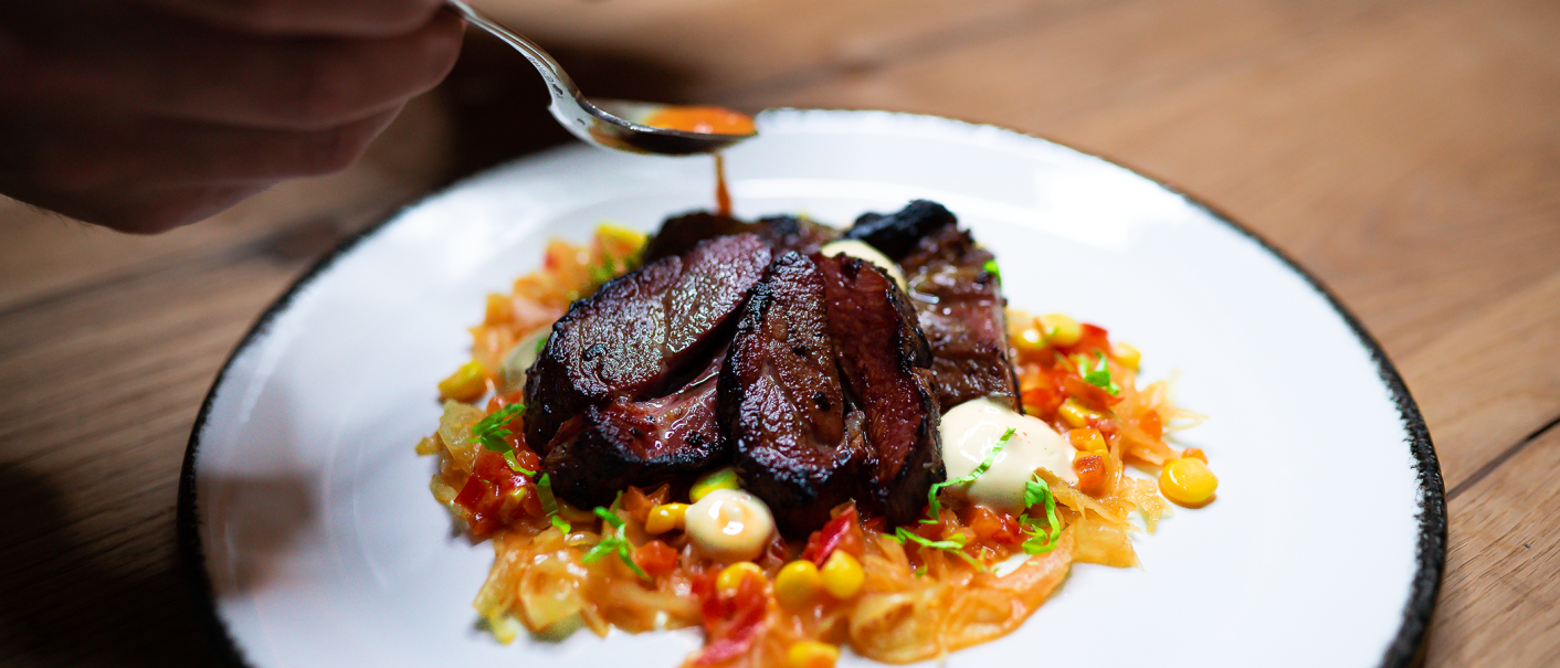 An artfully arranged dish with meat, vegetables and sauce on a white plate. One hand holds a spoon over the plate., © Cara Schmid An artfully arranged dish with meat, vegetables and sauce on a white plate. One hand holds a spoon over the plate., © Cara Schmid