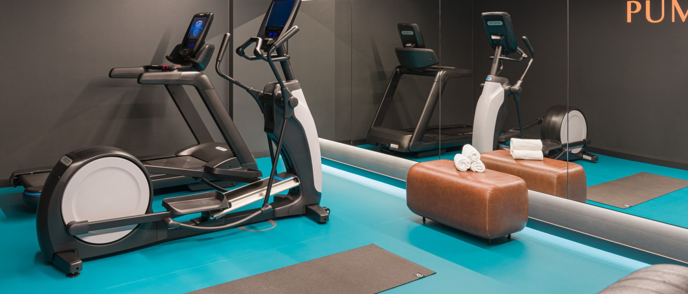 Modernes Fitnessstudio mit Laufband, Crosstrainer, Spiegelwand und blauer Bodenmatte. Ein brauner Hocker mit Handtüchern steht bereit., © EmiLu Hotel GmbH
