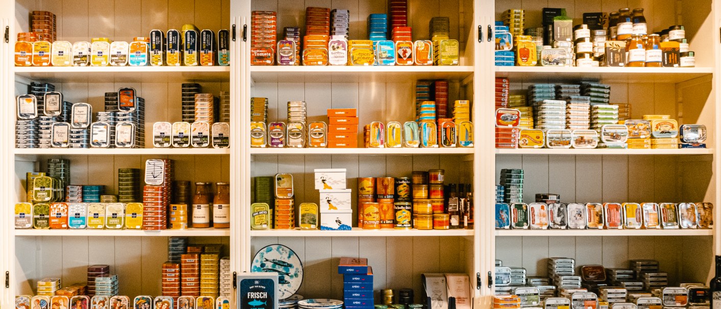Ein Regal in einer Weinbar, gefüllt mit bunten Dosen und Feinkostprodukten. Oben stehen dekorative Körbe., © SMG, Sarah Schmid